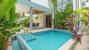 Ein moderner Poolbereich mit einem klaren, blauen Pool, umgeben von tropischer Vegetation und einer überdachten Sitzgelegenheit.