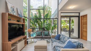 Ein modernes Wohnzimmer mit großem Fernseher, bequemen Sitzgelegenheiten und Blick auf Pool und Grünfläche.