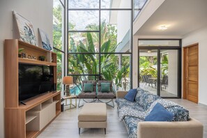 A modern living room with a large TV, comfortable seating, and a view of a pool and greenery. - Tropical Oasis 3-Min Walk to Playa Pelada Beach (Nosara)