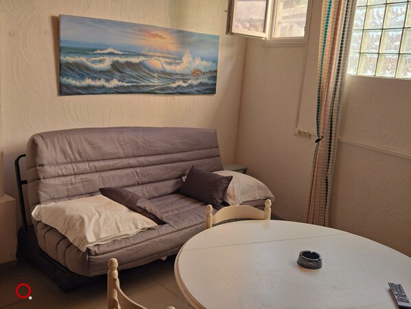 Une pièce comprenant un canapé, une table, un tableau représentant l’océan et une fenêtre avec des rideaux.