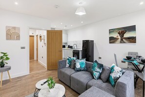 A modern living room with a grey sofa, a dining table, a kitchen area, and a hallway leading to another room. - Central | FREE parking | Connections to Manchester (Cheshire)