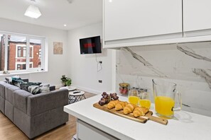 A modern kitchen with a marble countertop, a TV mounted above it, and a dining area with a grey sofa and a small table. - Central | FREE parking | Connections to Manchester (Cheshire)