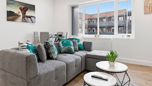 A modern living room with a grey sectional sofa, a small round table, a remote control, and a window with a view of buildings. - Central | FREE parking | Connections to Manchester (Cheshire)