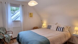 Ein Schlafzimmer mit Bett, Sessel, einem Fenster mit Vorhängen, einer Lampe und einem Wandbild.