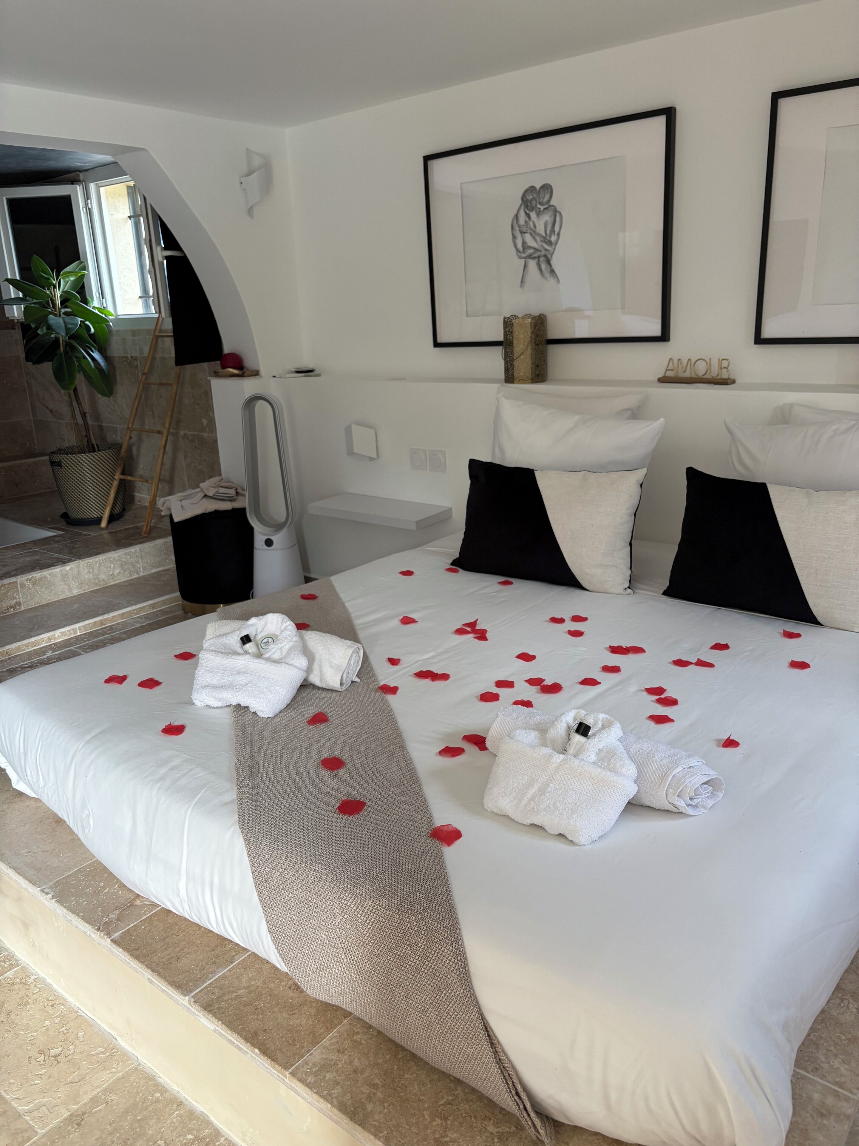 Ett hotellrum med en stor säng, vita sängkläder, en handduksställning, ett litet bord, en växt och inramade konstverk på väggen.