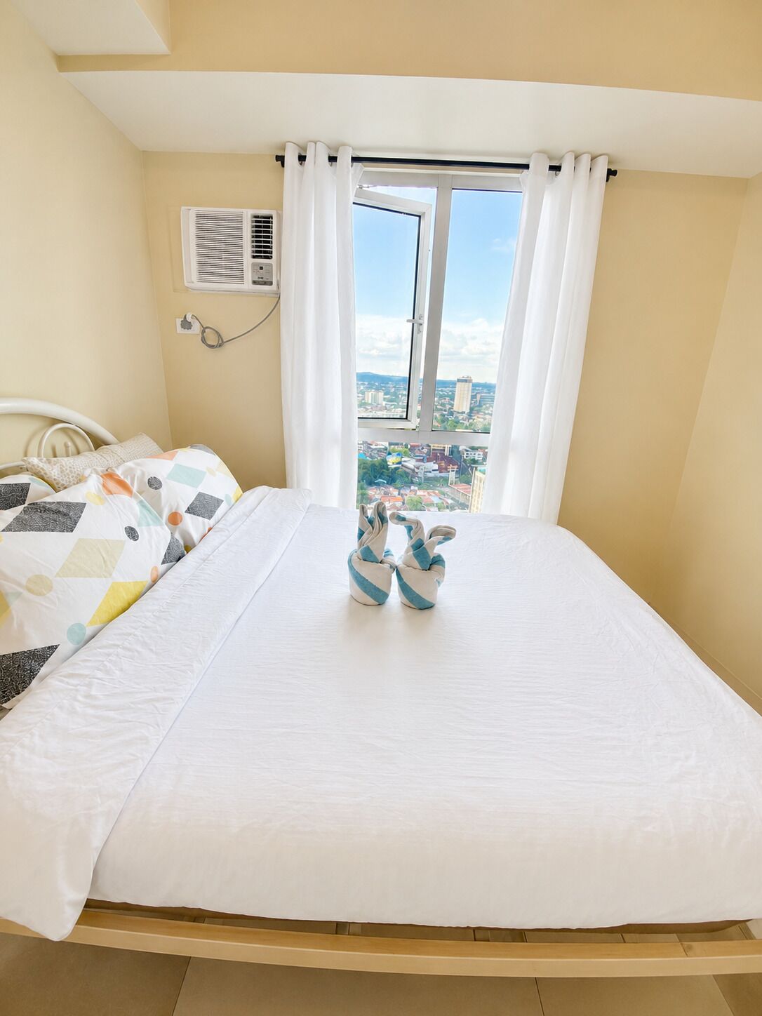 A bedroom with a bed, a window with curtains, an air conditioning unit, and a view of a cityscape.