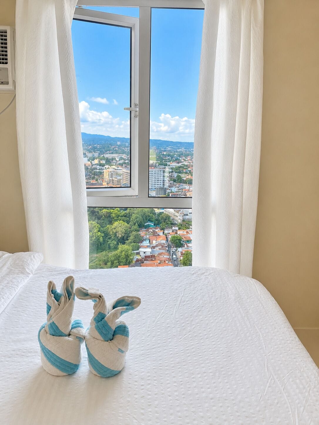Una cama con vistas a un paisaje urbano a través de una ventana con cortinas blancas.