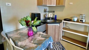 A compact kitchen with wooden cabinets, a dining table with chairs, a coffee machine, and a small refrigerator.