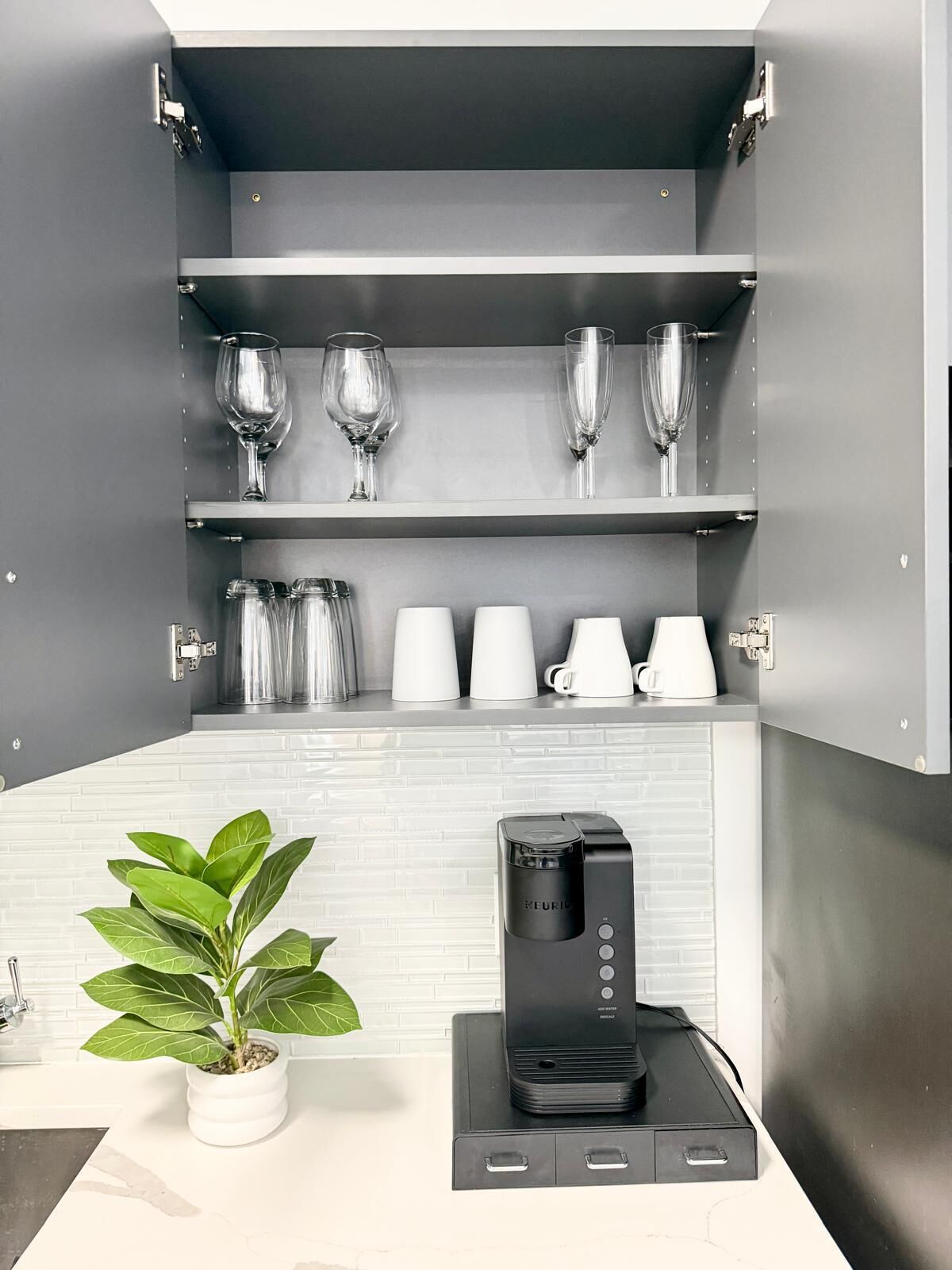 Een keuken met een koffiezetapparaat, een plant in pot en planken met glazen en kopjes.