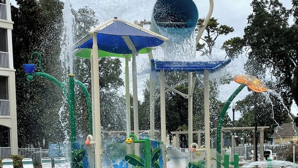 Estructura acuática con múltiples chorros de agua y un tobogán, rodeada por una piscina.