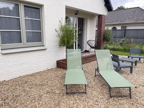 Una casa con fachada blanca, una chimenea de ladrillo y un patio de grava con dos sillones verdes y una mesita.