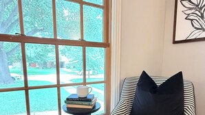 A room with a large window, a striped chair, a small table with books and a cup, and a framed artwork on the wall. - Hilltop Hideaway Near TCU, Dickies Arena & Medical District (Fort Worth)