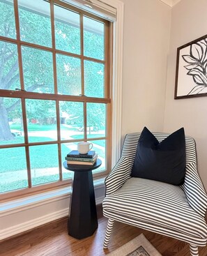 Ein Zimmer mit einem großen Fenster, einem gestreiften Sessel, einem kleinen Tisch mit Büchern und einer Tasse sowie einem gerahmten Kunstwerk an der Wand.