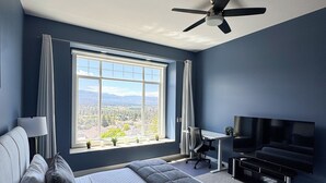 A modern bedroom with a large bed, a desk, a TV, and a view of the outdoors. - Beautiful Mountain & Lake Views/Upper Level Home/King Bed/BBQ/Sleeps 5 (Kelowna)