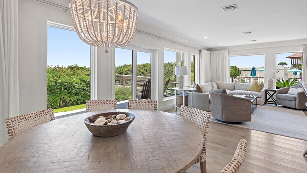 A spacious living room with a large chandelier, a round dining table with a bowl, and a view of the outdoors through large windows. - Palms of Dune Allen 104 (Santa Rosa Beach)