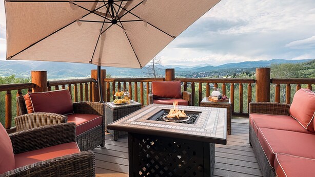 A deck with wicker furniture, a fire pit, and a table with a bottle and grapes. - Log Home w/ Epic Views, Hot Tub | Starfall Summit (Steamboat Springs)