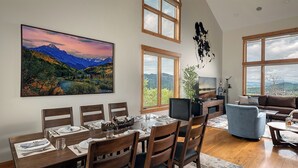 A dining area with a table set for four, a sofa, and a TV in a room with large windows and a mountain view. - Log Home w/ Epic Views, Hot Tub | Starfall Summit (Steamboat Springs)