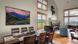 Ein Essbereich mit Tisch für vier Personen, einer Couch und einem Fernseher in einem Zimmer mit großen Fenstern und Bergblick.