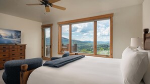 A bedroom with a large bed, a dresser, a chair, and a view of mountains through large windows. - Log Home w/ Epic Views, Hot Tub | Starfall Summit (Steamboat Springs)