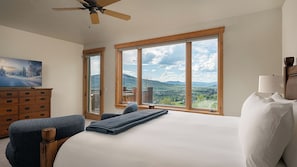 Ein Schlafzimmer mit einem großen Bett, einem Schminktisch, einem Sessel und Blick auf Berge durch große Fenster.