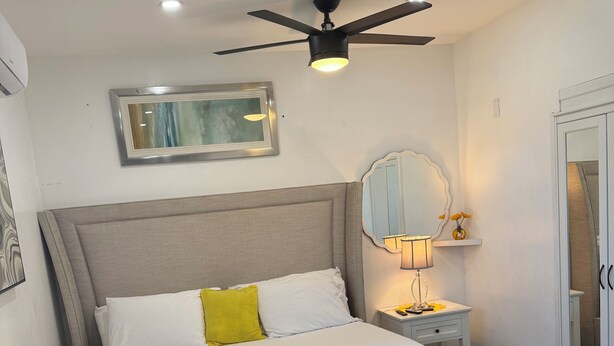 A neatly made bed with a white and yellow bedspread, a grey upholstered headboard, a ceiling fan, a bedside table with a lamp, and a mirror on the wall. - Private Cozy Studio (Sunrise)