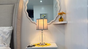 A bedroom with a bed, a bedside table with a lamp, a mirror, and a small shelf with flowers. - Private Cozy Studio (Sunrise)
