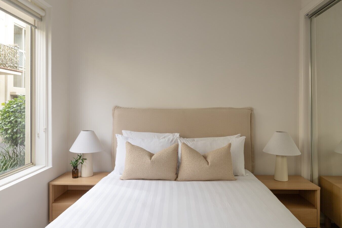 A bedroom with a bed, two bedside tables, two lamps, a window, and a view of greenery outside.