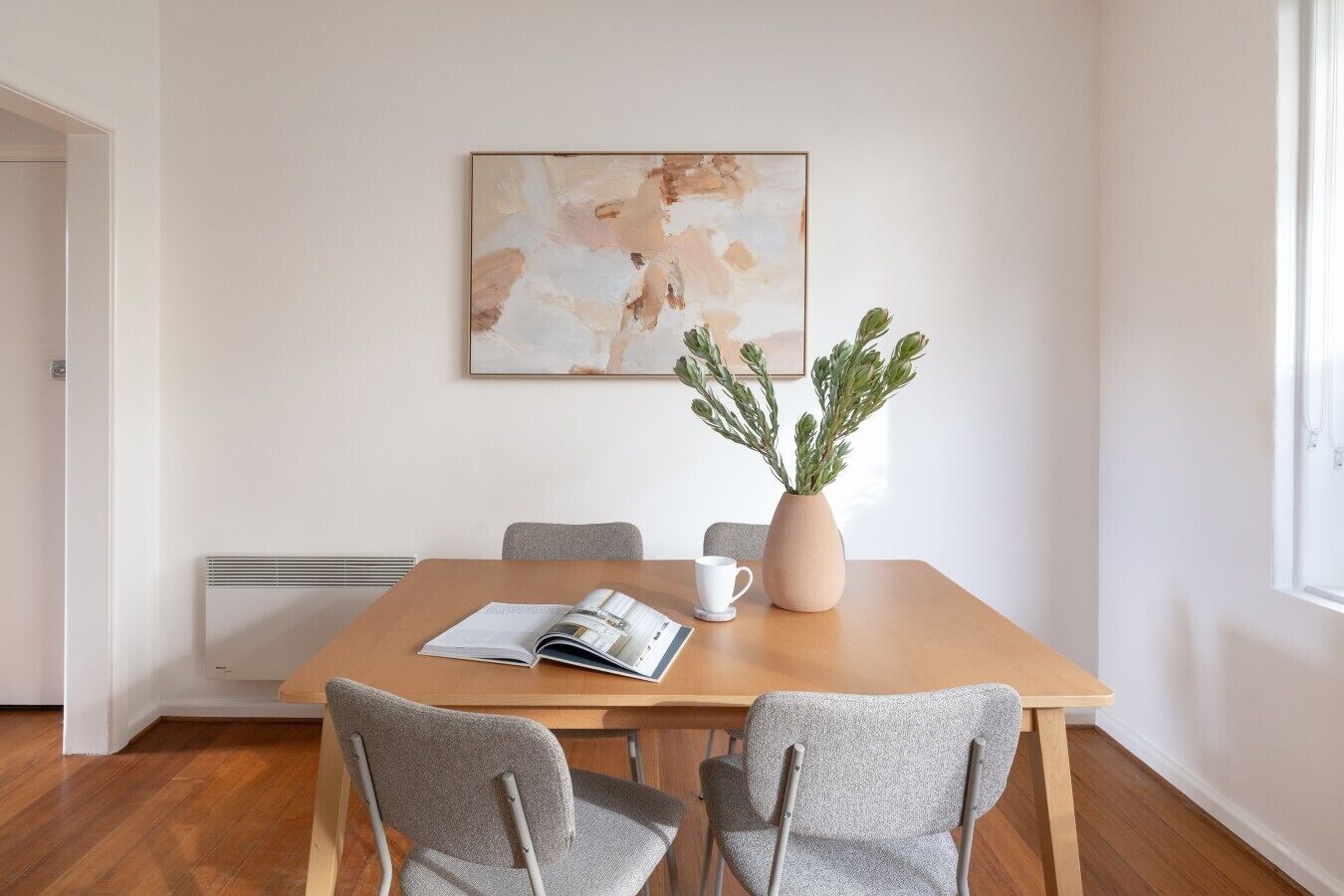 Ein Essbereich mit einem Holztisch, zwei grauen Stühlen, einer Vase mit Grünpflanzen, einer Zeitschrift und einem gerahmten abstrakten Gemälde an der Wand.