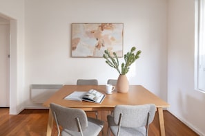 A dining area with a wooden table, two grey chairs, a vase with green plants, a magazine, and a framed abstract painting on the wall. - Beautifully Styled Apartment in St Kilda East (Saint Kilda East)