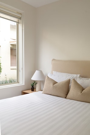 A bedroom with a bed, a bedside lamp, and a window. - Beautifully Styled Apartment in St Kilda East (Saint Kilda East)