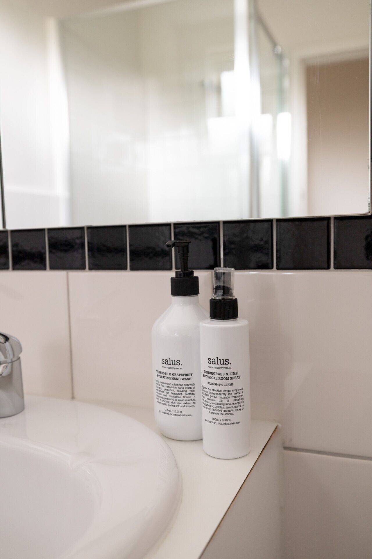 A bathroom with a white sink, two bottles of salus botanical skincare products, and a black and white tiled backsplash.