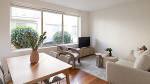 Ein modernes Wohnzimmer mit einem Holztisch, zwei Stühlen, einer Couch, einem Fernseher und einem großen Fenster mit Blick auf Grünflächen und Gebäude.