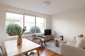 A modern living room with a wooden table, two chairs, a sofa, a television, and a large window with a view of greenery and buildings. - Beautifully Styled Apartment in St Kilda East (Saint Kilda East)