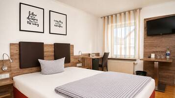 A hotel room with a bed, a desk, a chair, a TV, and two motivational posters on the wall.