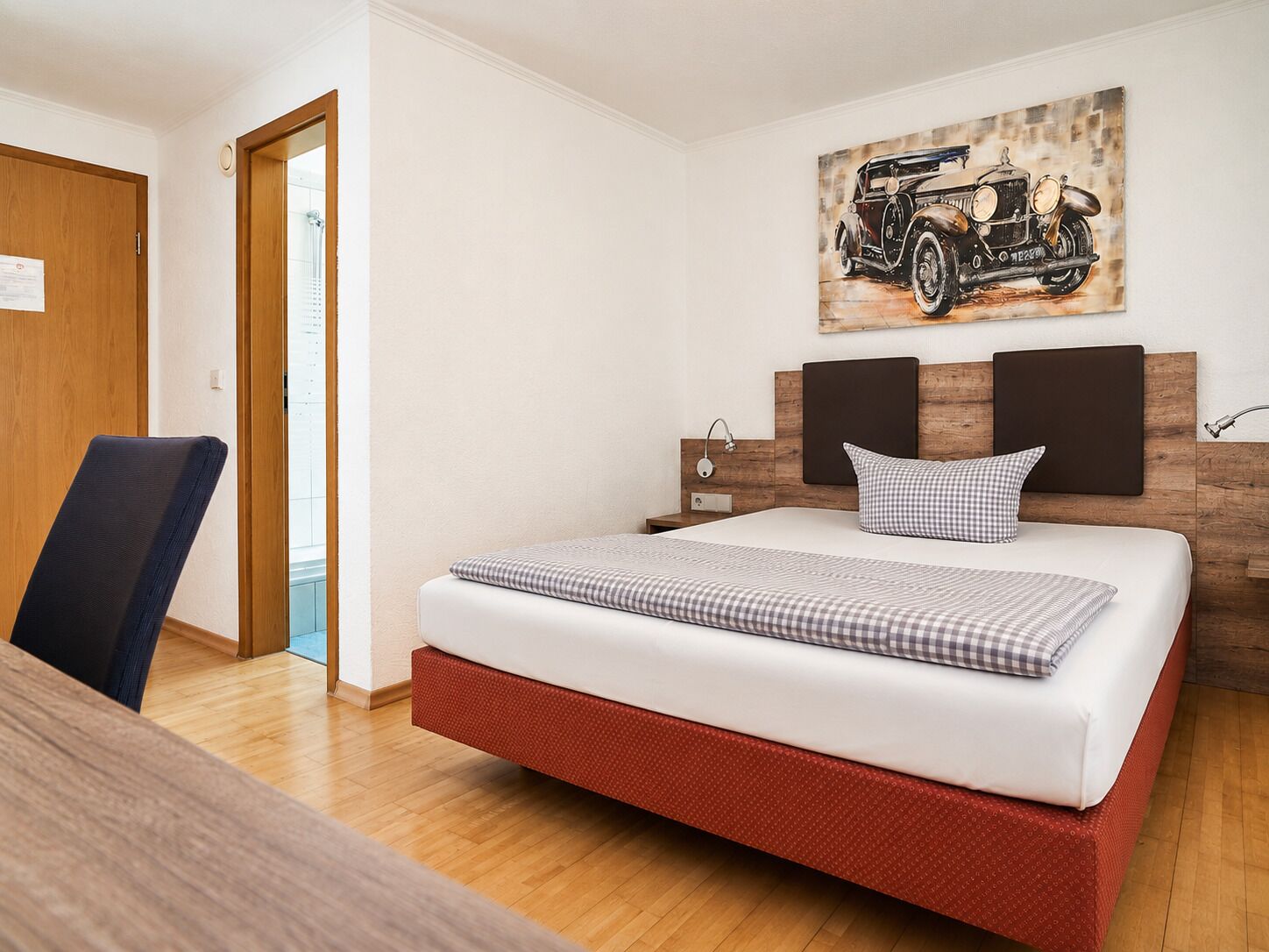 A bedroom with a bed, a chair, a wooden desk, a painting of a car, and a wooden door.