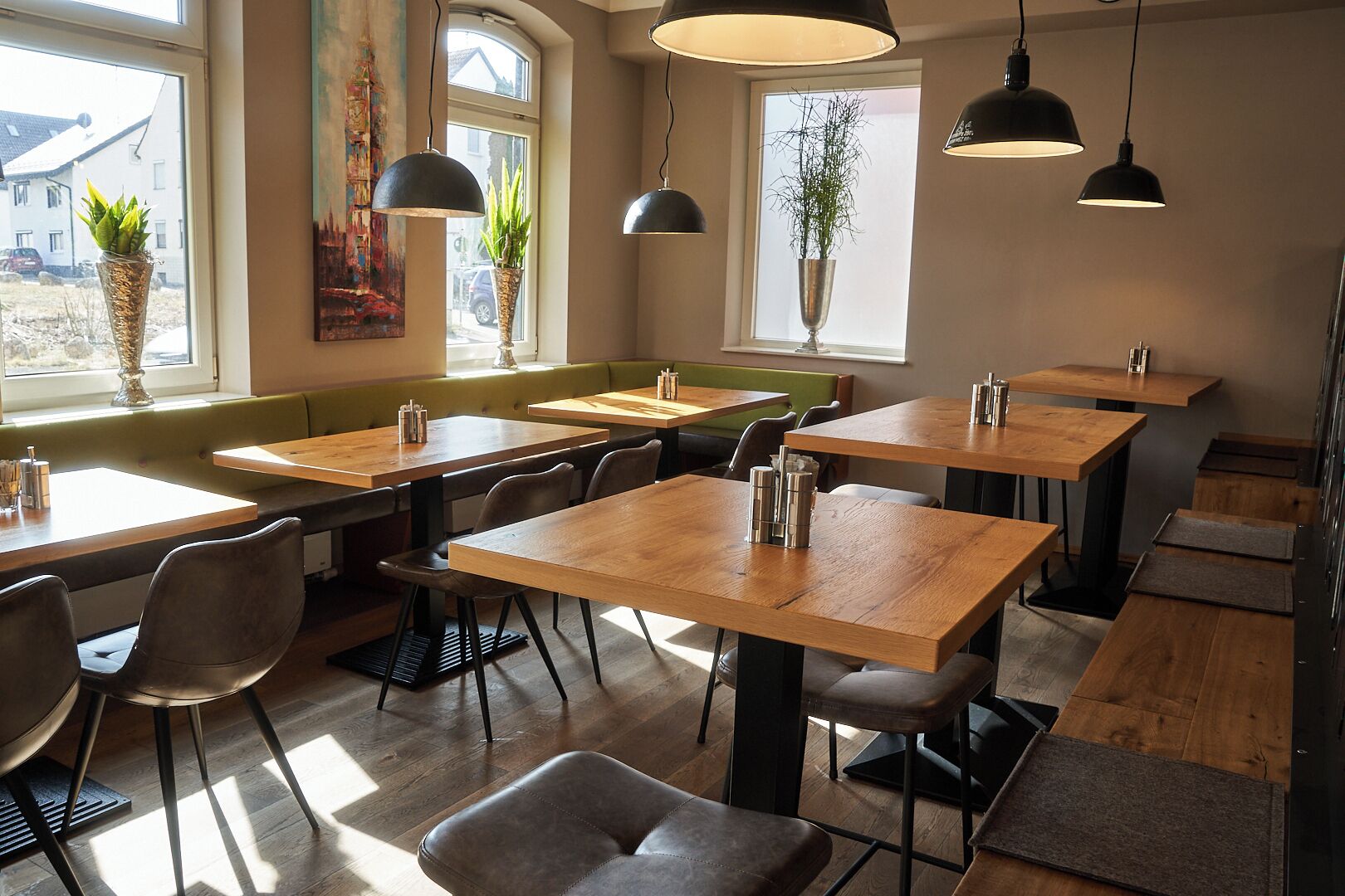 A restaurant interior with wooden tables and chairs, black pendant lights, and potted plants.