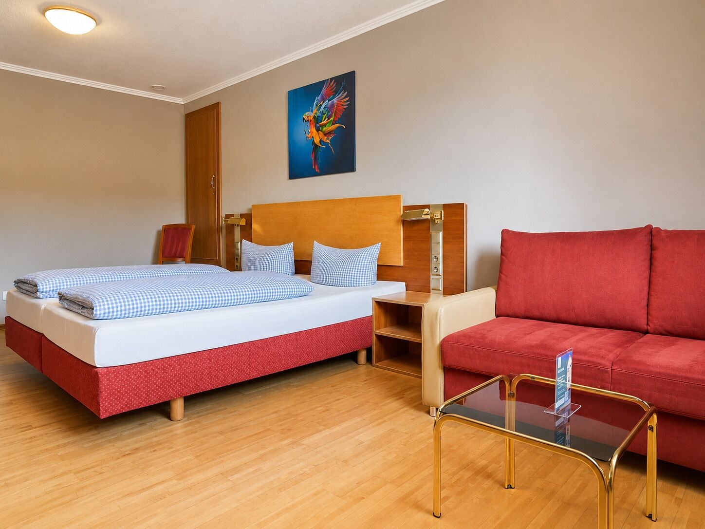 A hotel room with a bed, a red sofa, a glass coffee table, and a painting on the wall.