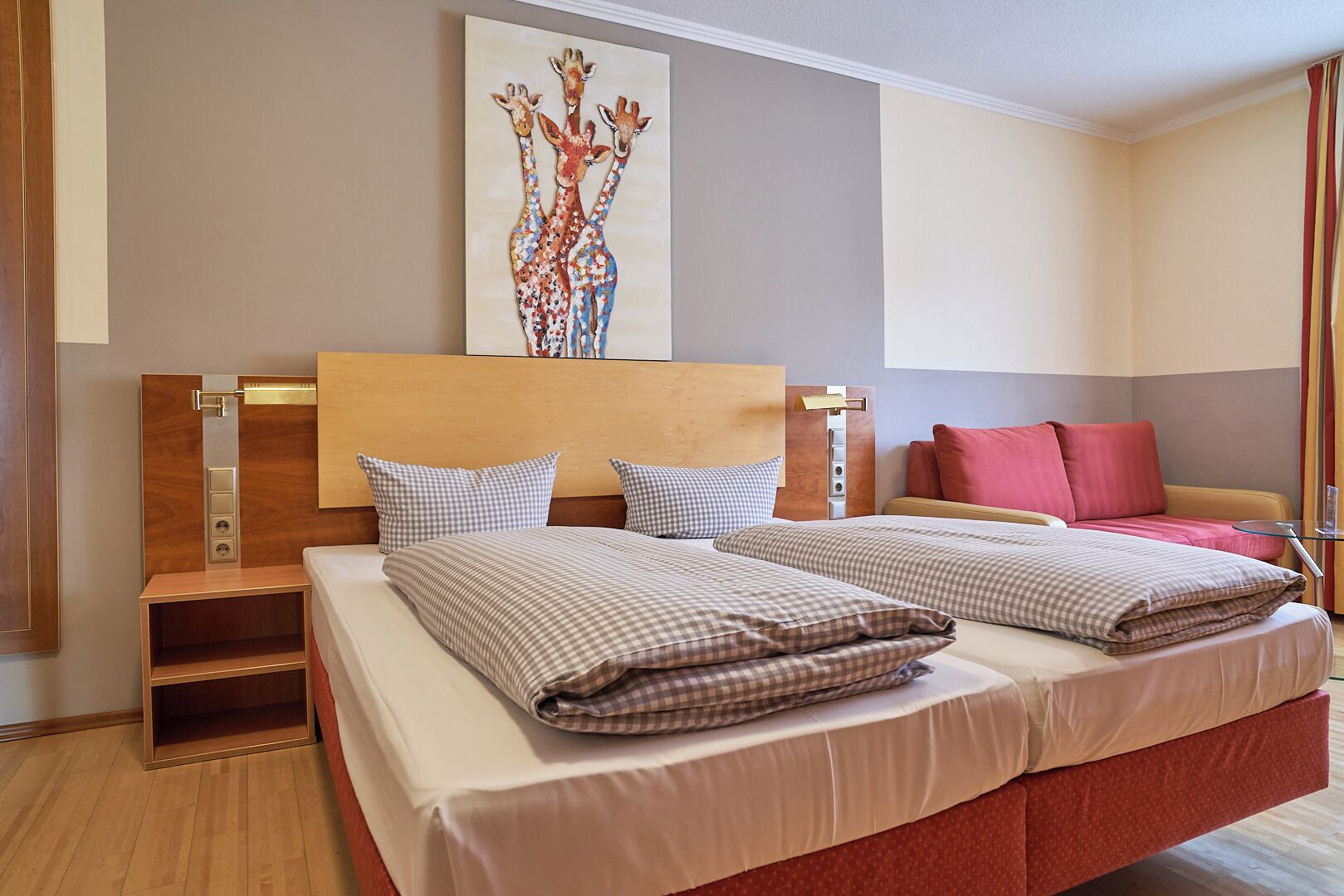 Quarto de hotel com duas camas, uma cabeceira de madeira, uma obra de arte na parede e um sofá vermelho.