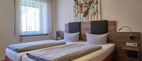 Cama doble con ropa de cama a cuadros, cabecero con una obra de arte de cabeza de ciervo, mesita de noche con una lámpara y ventana con cortinas translúcidas.