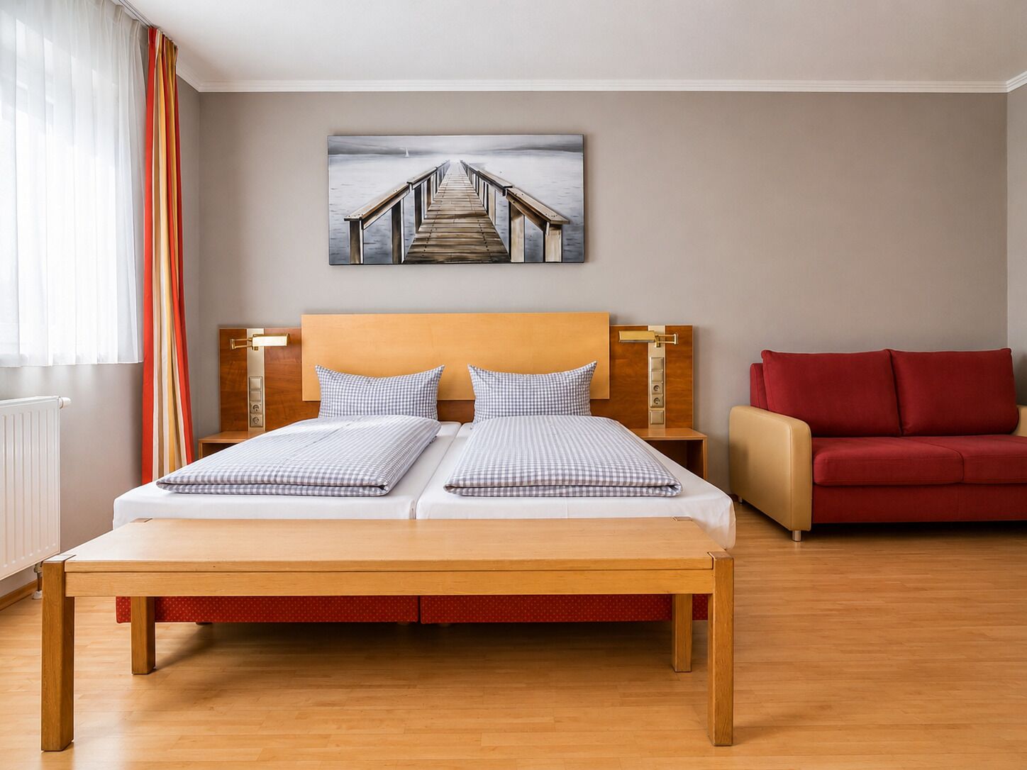 A hotel room with a wooden bed, a red sofa, and a painting of a pier on the wall.
