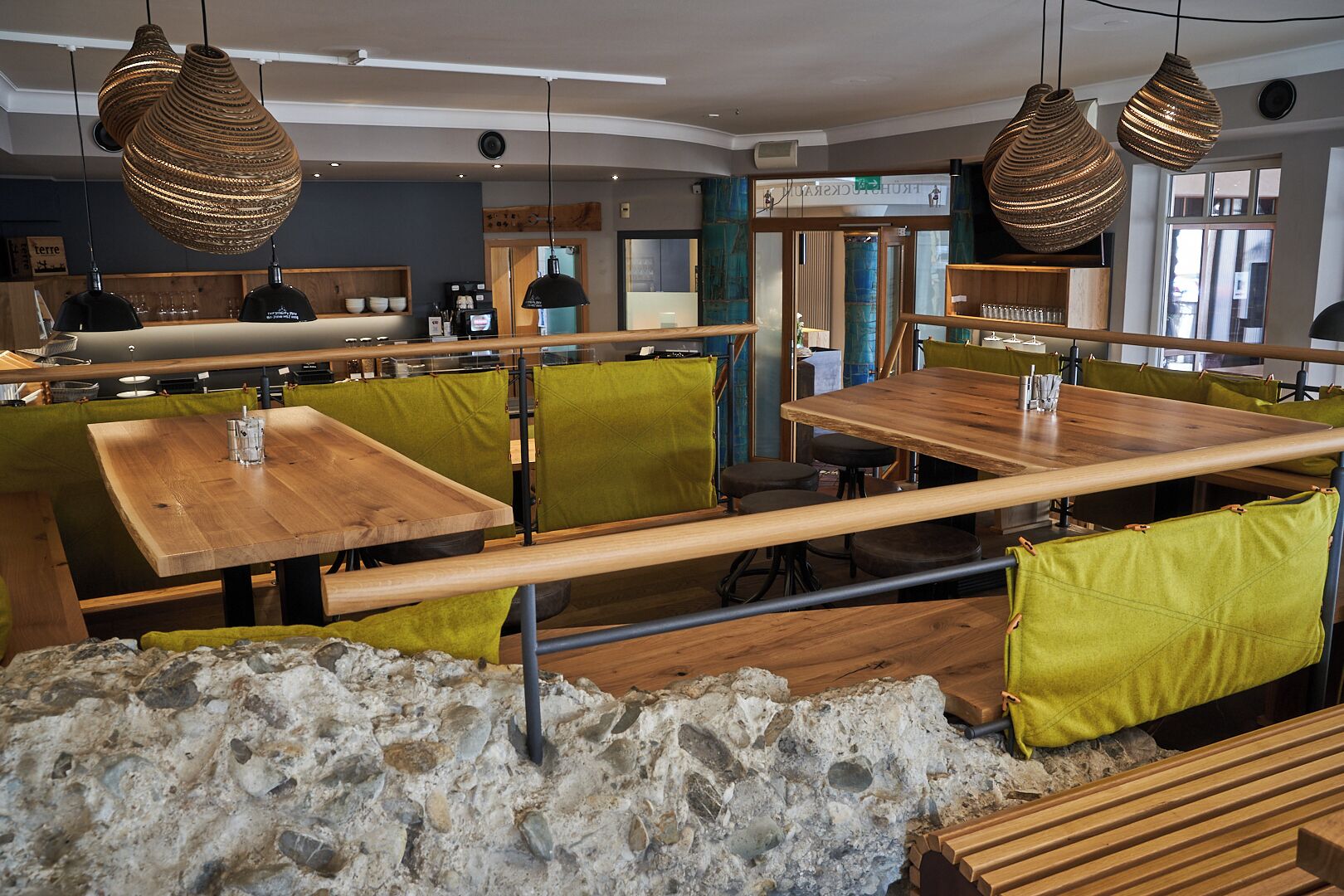 Ein Restaurantinneres mit Holztischen, grün gepolsterten Sitzplätzen, hängenden gewebten Lampen und einer Steinwand.