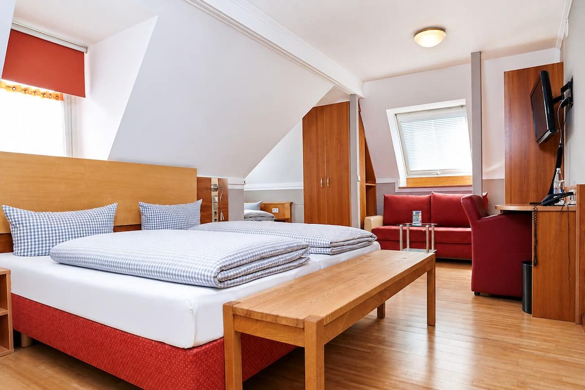 A bedroom with a bed, a wooden bench, a red sofa, a desk with a phone, and a TV mounted on the wall.