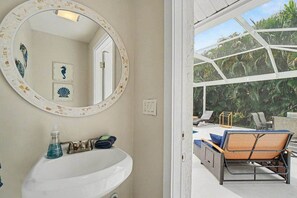 Ein Badezimmer mit rundem Spiegel, Waschbecken und Pflegeprodukten. Es gibt einen Zugang zu einem Balkon mit Gartenmöbeln und Blick ins Grüne.