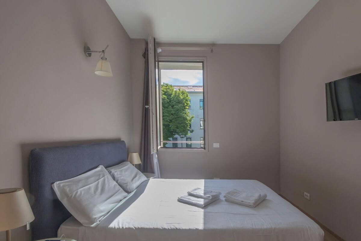 Una habitación de hotel con una cama, un televisor, un ventanal con vistas y una lámpara.