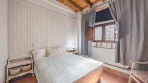 A bedroom with a wooden ceiling, a bed with a patterned bedspread, a nightstand, a chair, and a window with curtains. - Holiday Home 'El Mimbral' with Shared Pool, Wi-Fi and Air Conditioning (Vejer de la Frontera)