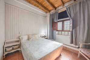 Ein Schlafzimmer mit Holzboden, einem Bett mit gemustertem Bettbezug, einem Nachttisch, einem Sessel und einem Fenster mit Vorhängen.