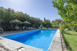 Una piscina rectangular amb un mur de pedra, envoltada de gandules i paraigües de palla, situada en un jardí exuberant.