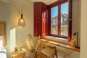 A room with a wooden desk, a chair, a lamp, a window with red shutters, and a view of buildings outside. - Porto Haven - Cedofeita 213 (Porto)