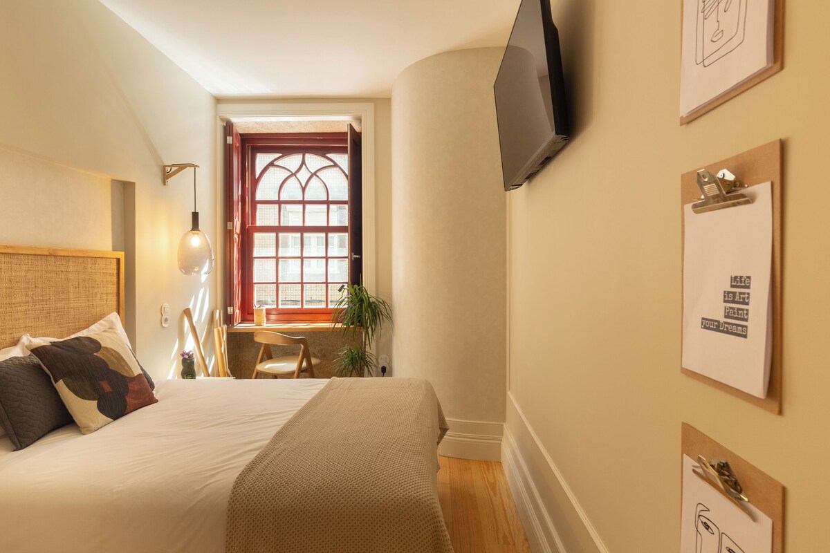 Quarto com cama, escrivaninha, cadeira, televisão e janela com vista.