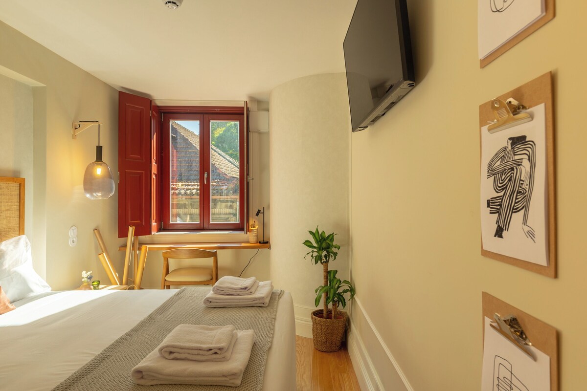 Quarto de hotel com uma cama, uma escrivaninha, uma televisão, uma janela com vista e obras de arte na parede.